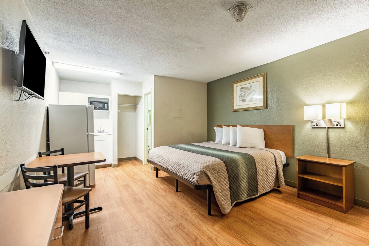 A hotel room with a bed, a desk, a chair, a TV, a lamp, and a shelf.