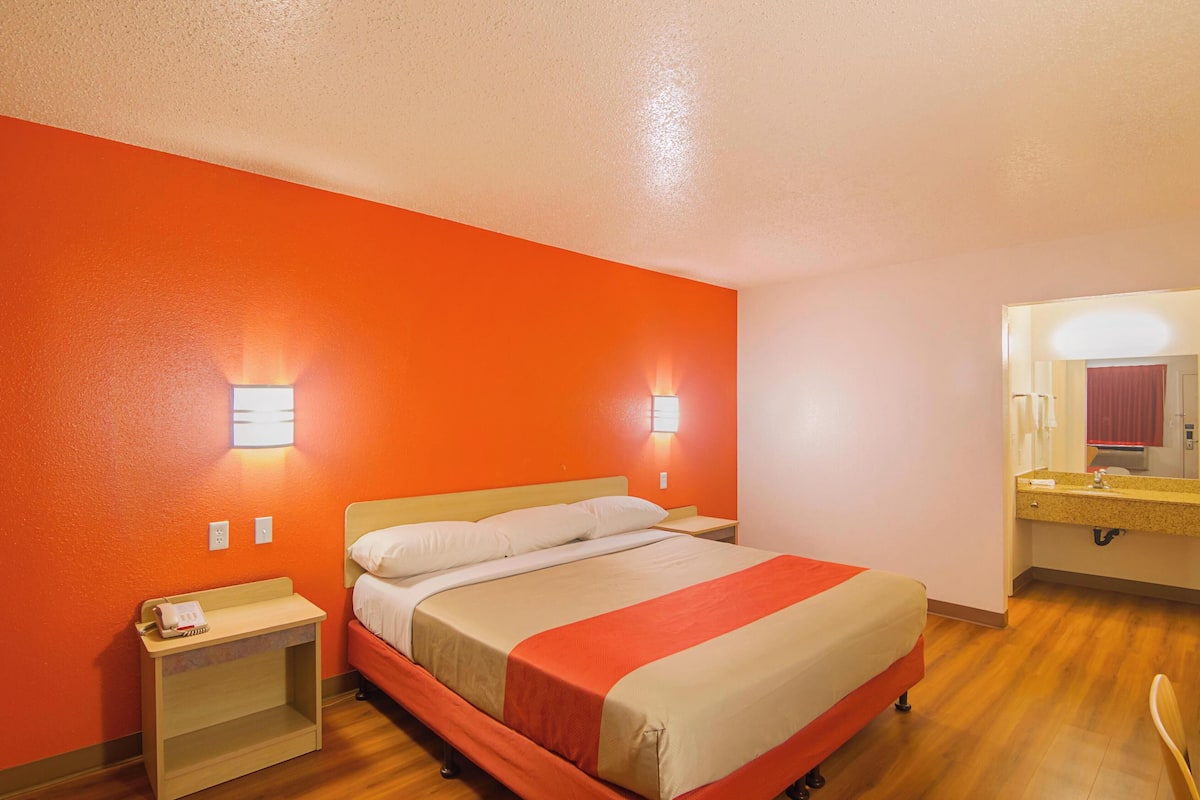 A hotel room with a single bed, a bedside table, a nightstand, a mirror, and a bathroom with a sink and mirror.