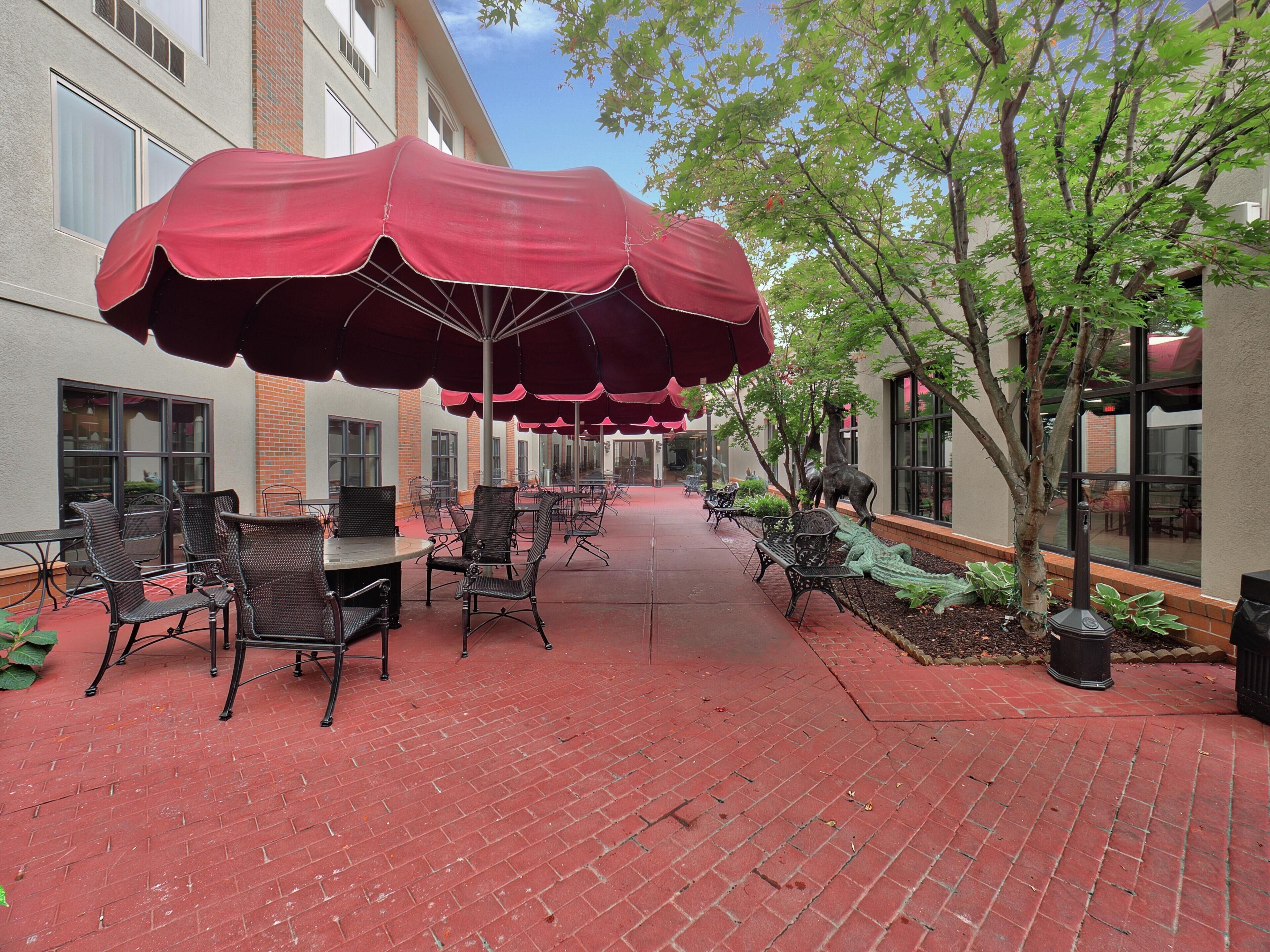 A brick-paved outdoor patio with tables and chairs under red umbrellas, surrounded by trees and a building with large windows.