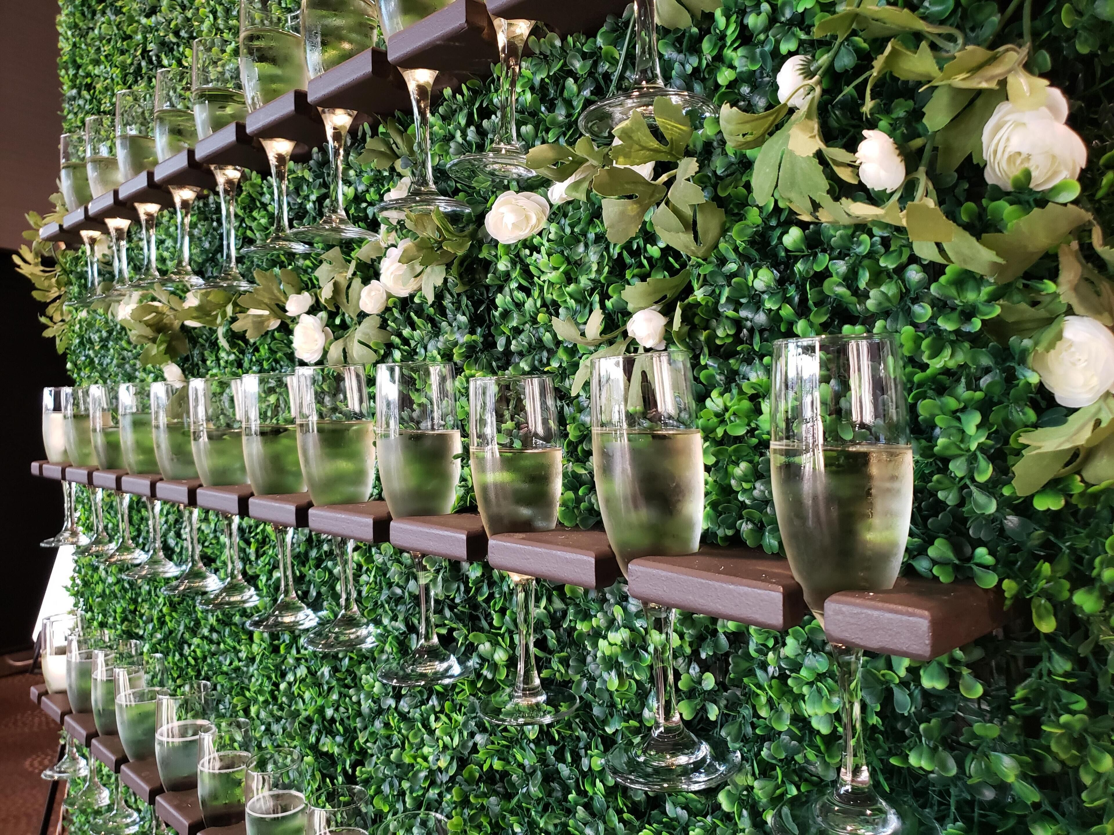 A wall of wine glasses arranged on shelves against a backdrop of green foliage and white flowers.