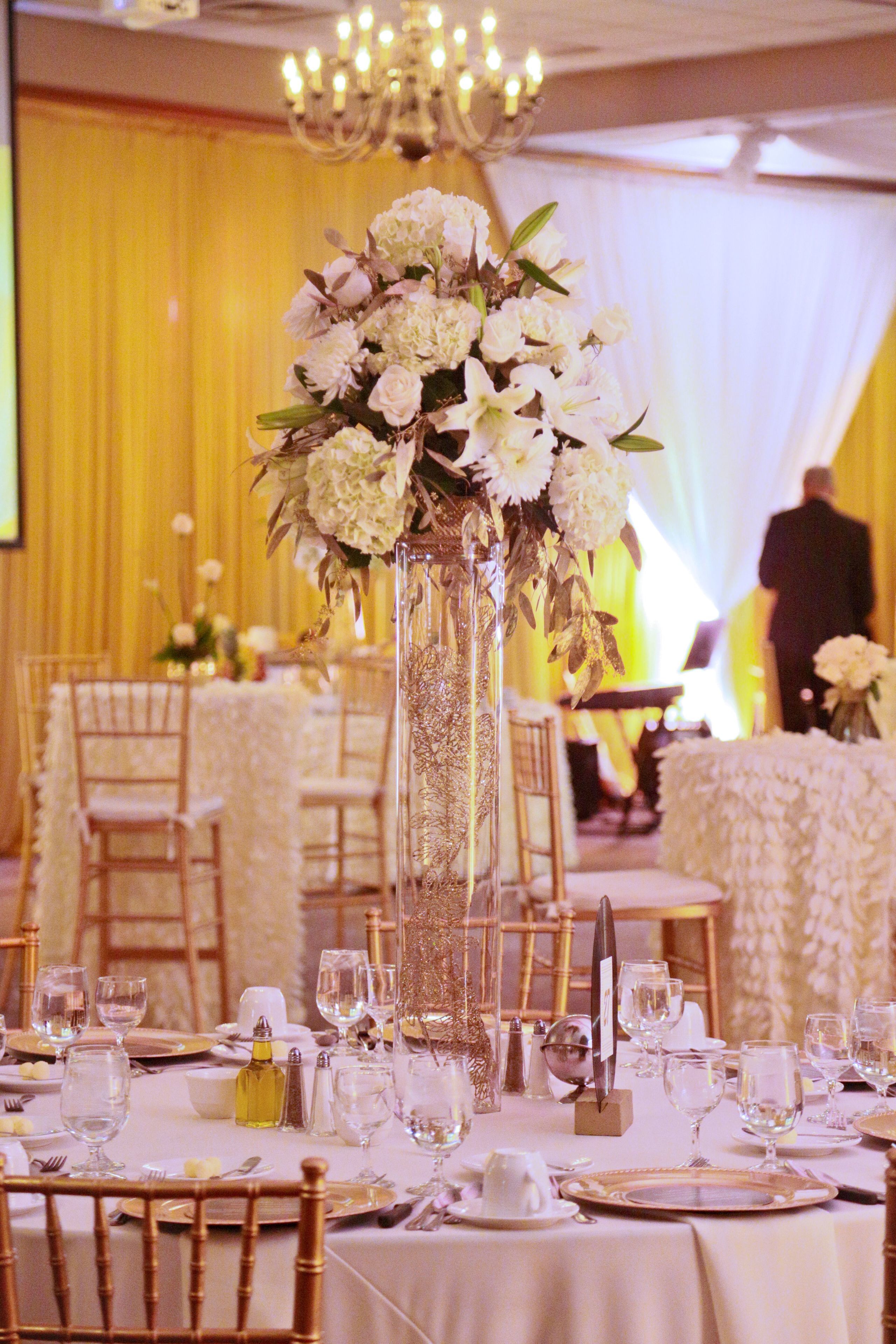 Salão de festas com mesas montadas para um evento formal, com vasos altos de flores, toalhas brancas e detalhes dourados.