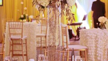Salão de festas com mesas montadas para um evento formal, com vasos altos de flores, toalhas brancas e detalhes dourados.