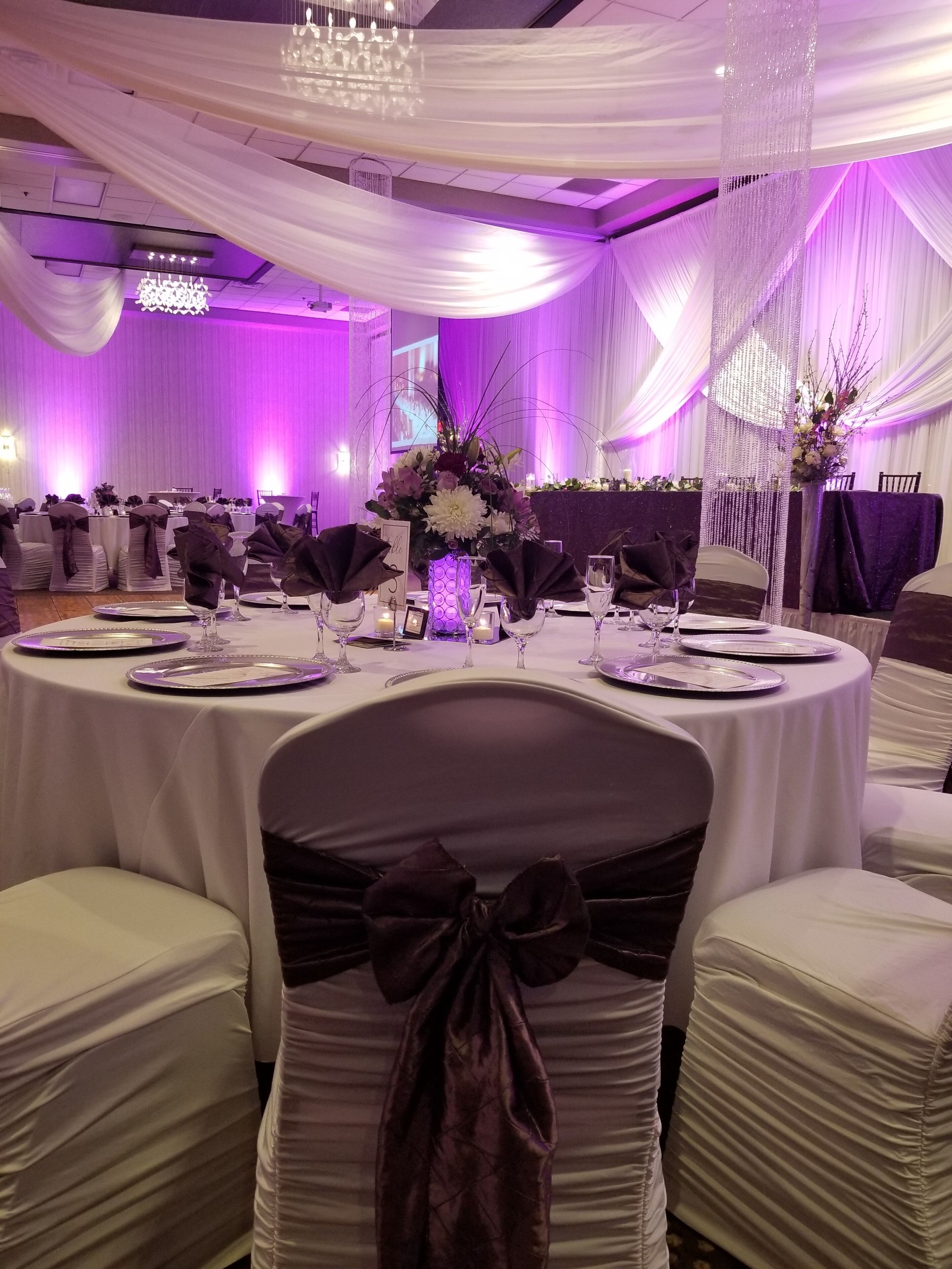 Un gran saló de banquets amb taules rodones preparades per a un esdeveniment formal, amb estovalls blancs, cadires fosques amb laços i centrals de flors elegants.
