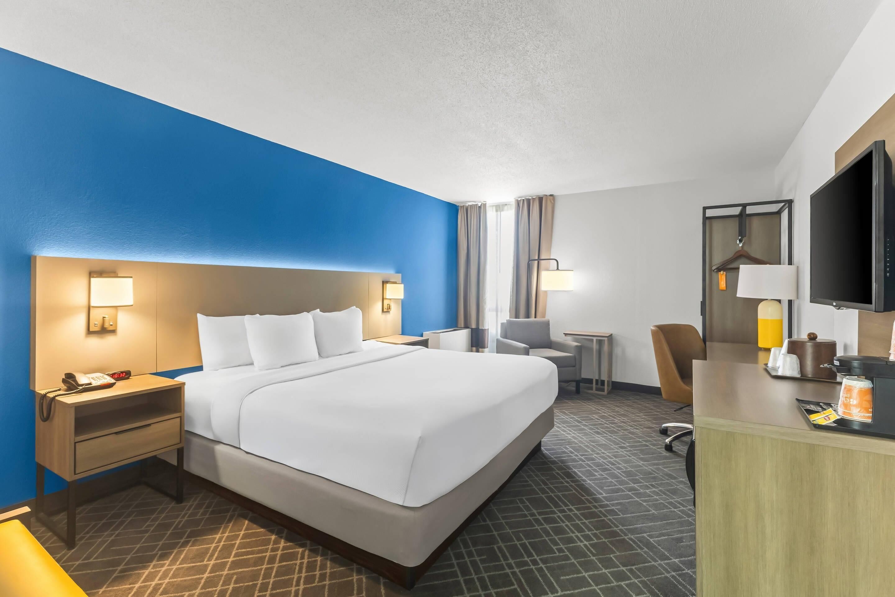 A hotel room with a large bed, bedside tables, a desk, a chair, a TV, and a minibar.