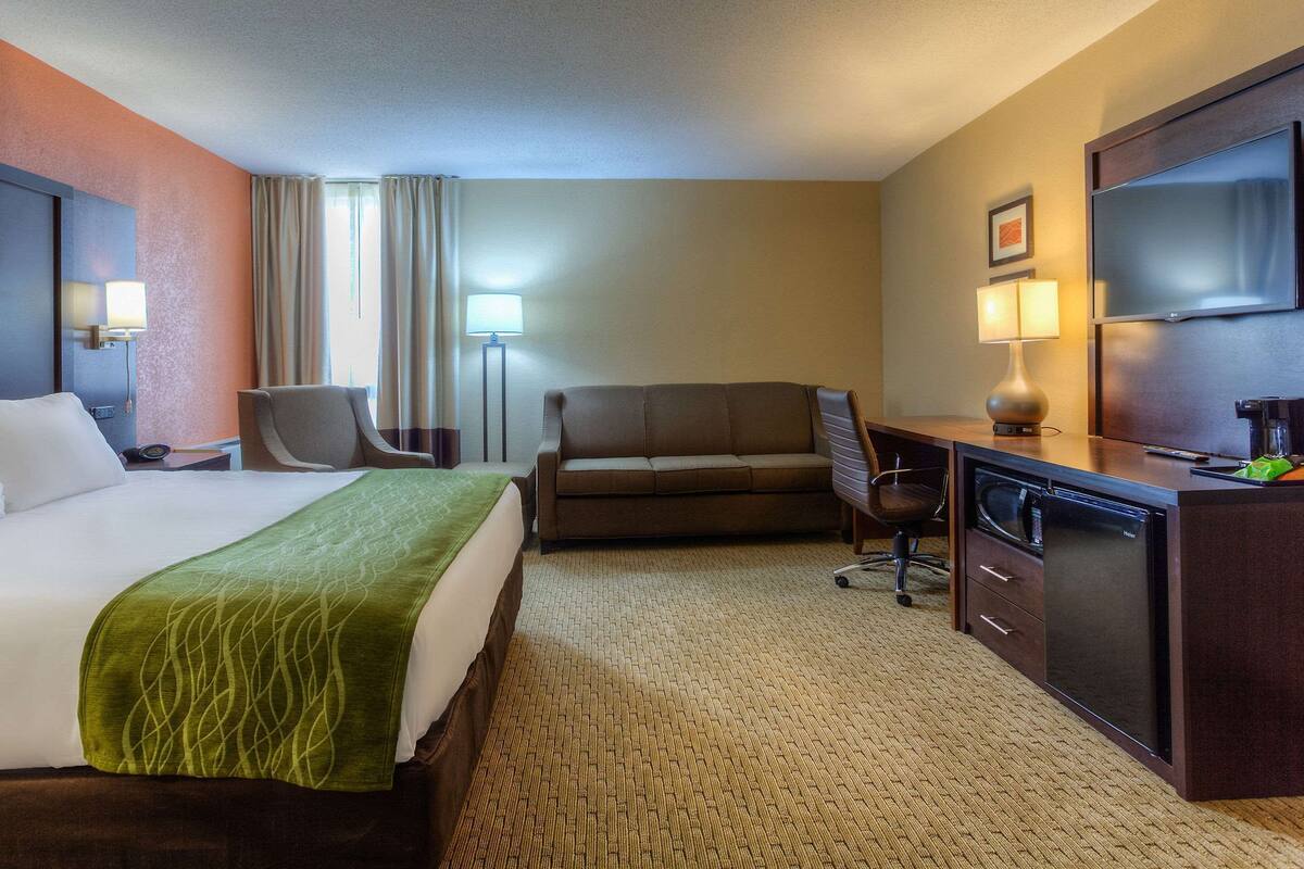 A hotel room with a bed, a sofa, a desk with a computer, a TV, and a lamp.