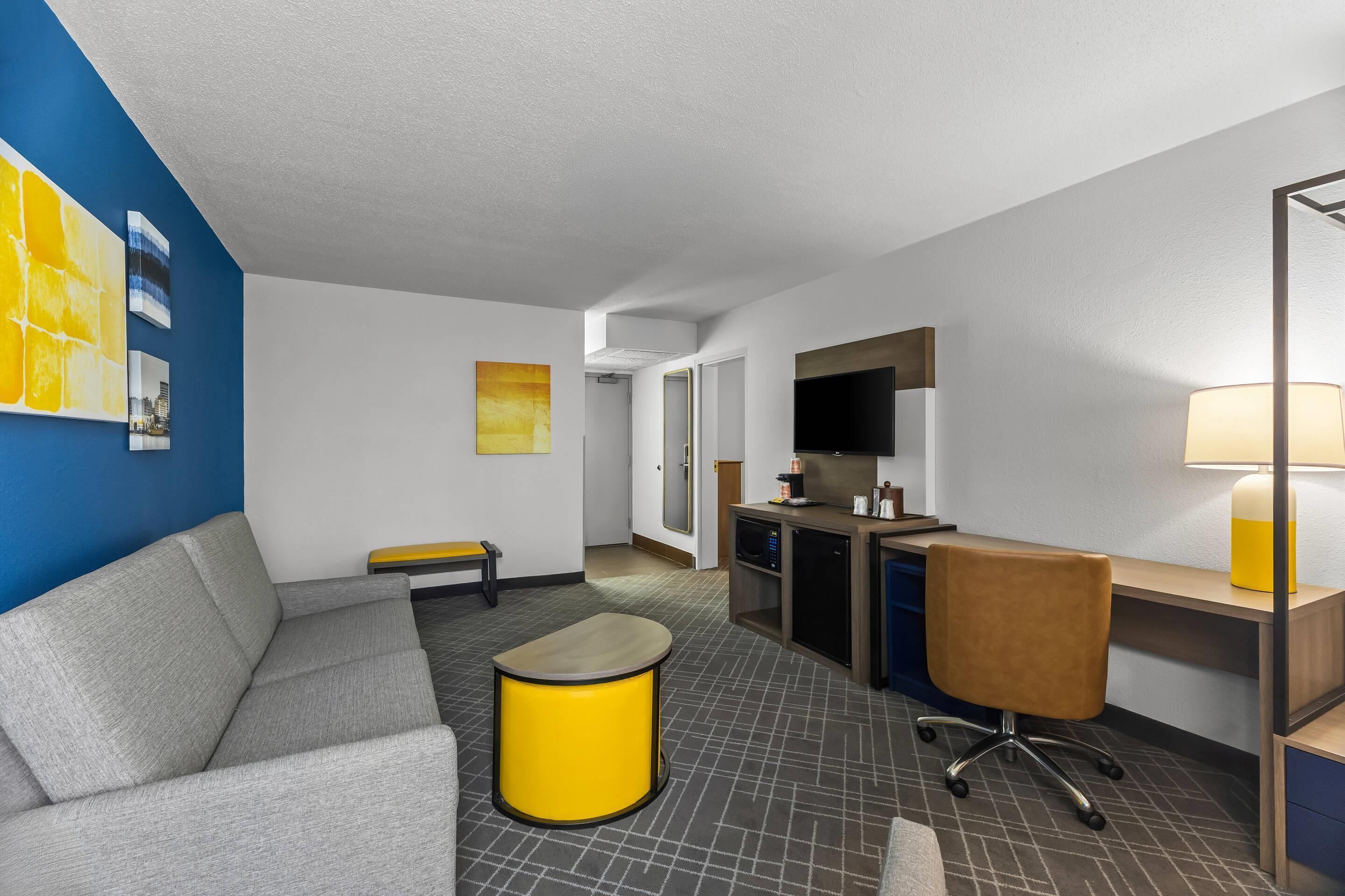 Hotellihuone, jossa on sohva, pieni pöytä, työpöytä tuolilla, mikroaaltouuni, kahvinkeitin ja taideteoksia seinillä.