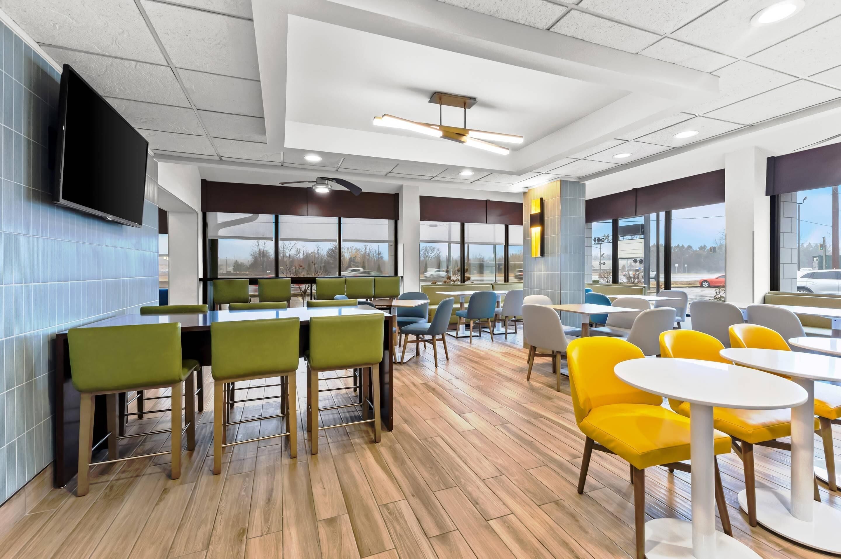 Une cafétéria moderne dotée de nombreuses tables et chaises, de grandes fenêtres et d’un téléviseur à écran plat fixé au mur.