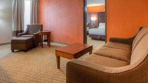 Una habitación de hotel con un sofá marrón, una silla, una mesita de madera y una cama con cabecero blanco.