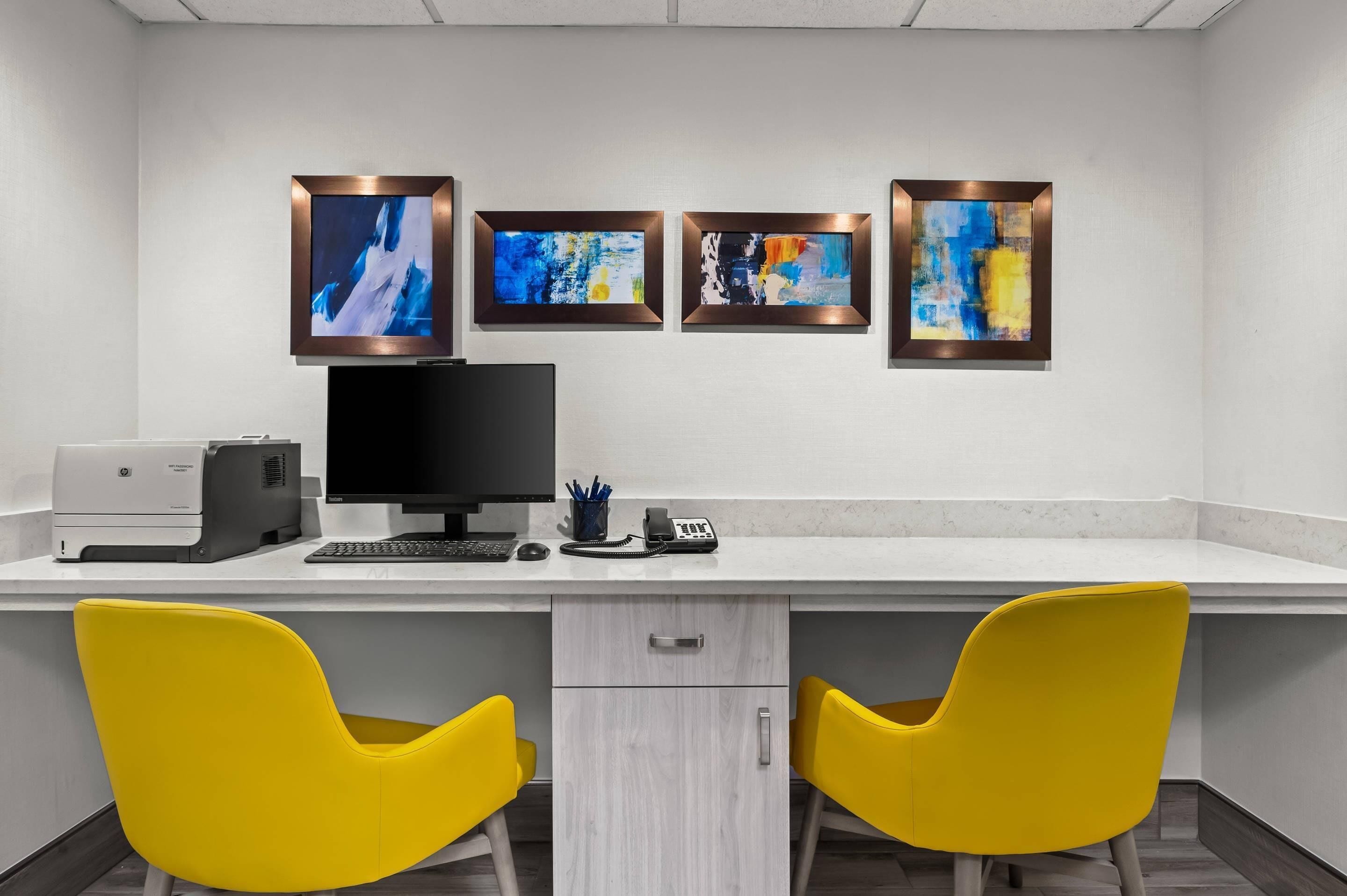 A modern office space with a desk, two yellow chairs, a computer, and four framed abstract paintings on the wall.
