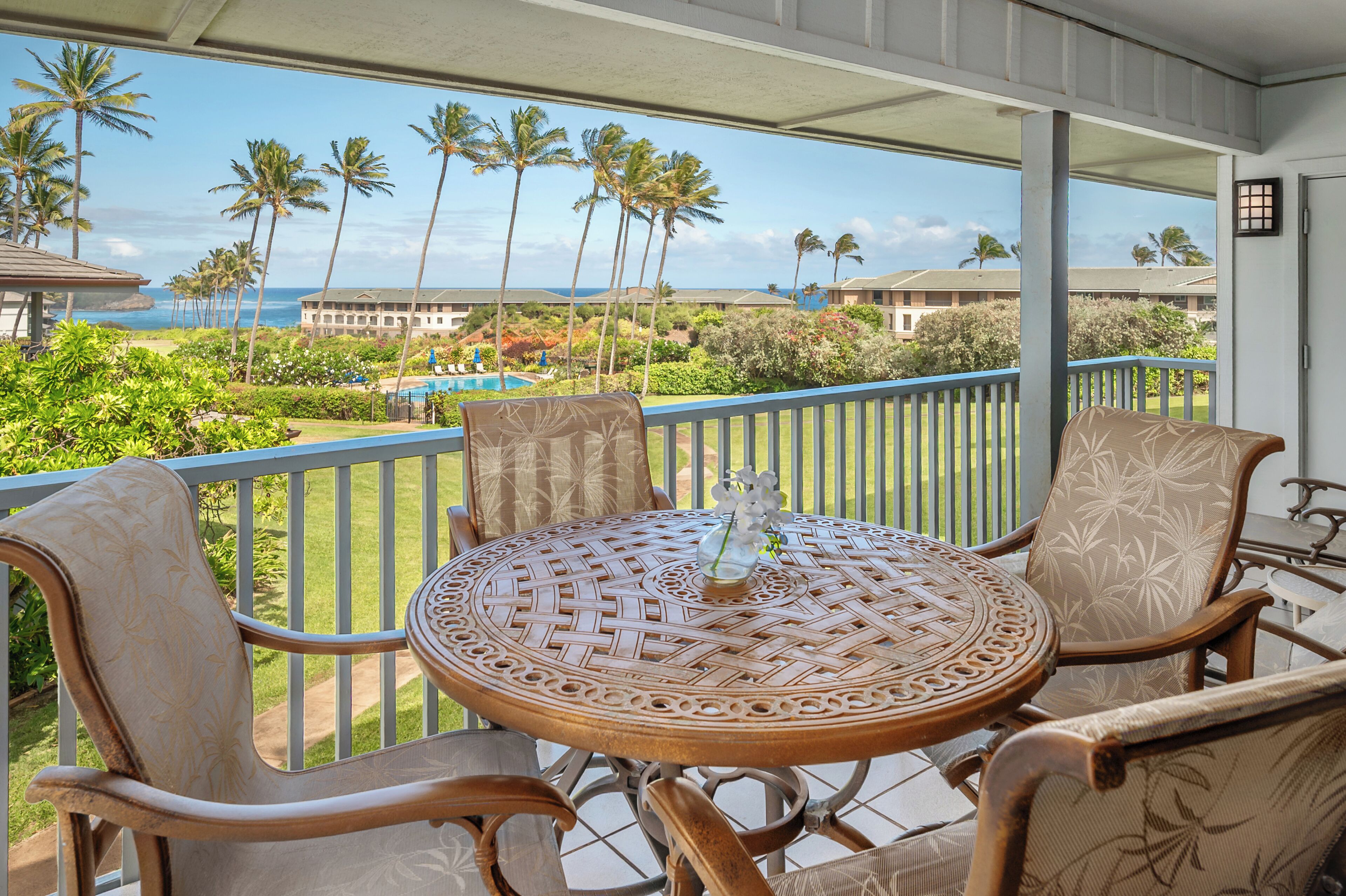 A balcony with wicker furniture, a small table with a vase, and a view of a resort with palm trees and a pool.