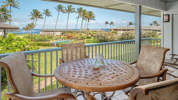 A balcony with wicker furniture, a small table with a vase, and a view of a resort with palm trees and a pool.