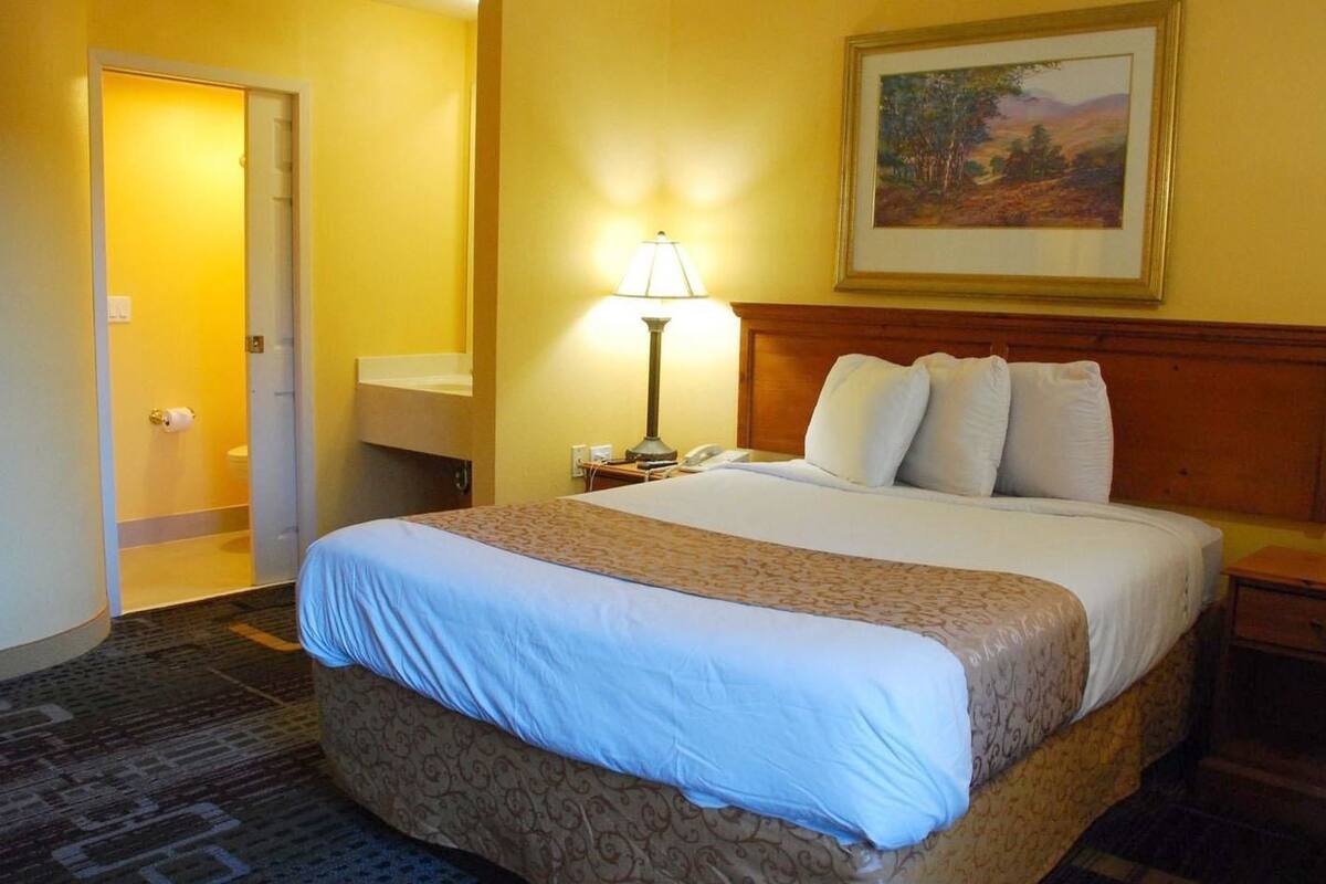 A hotel room with a large bed, a bedside lamp, a painting on the wall, and a bathroom door visible in the background.