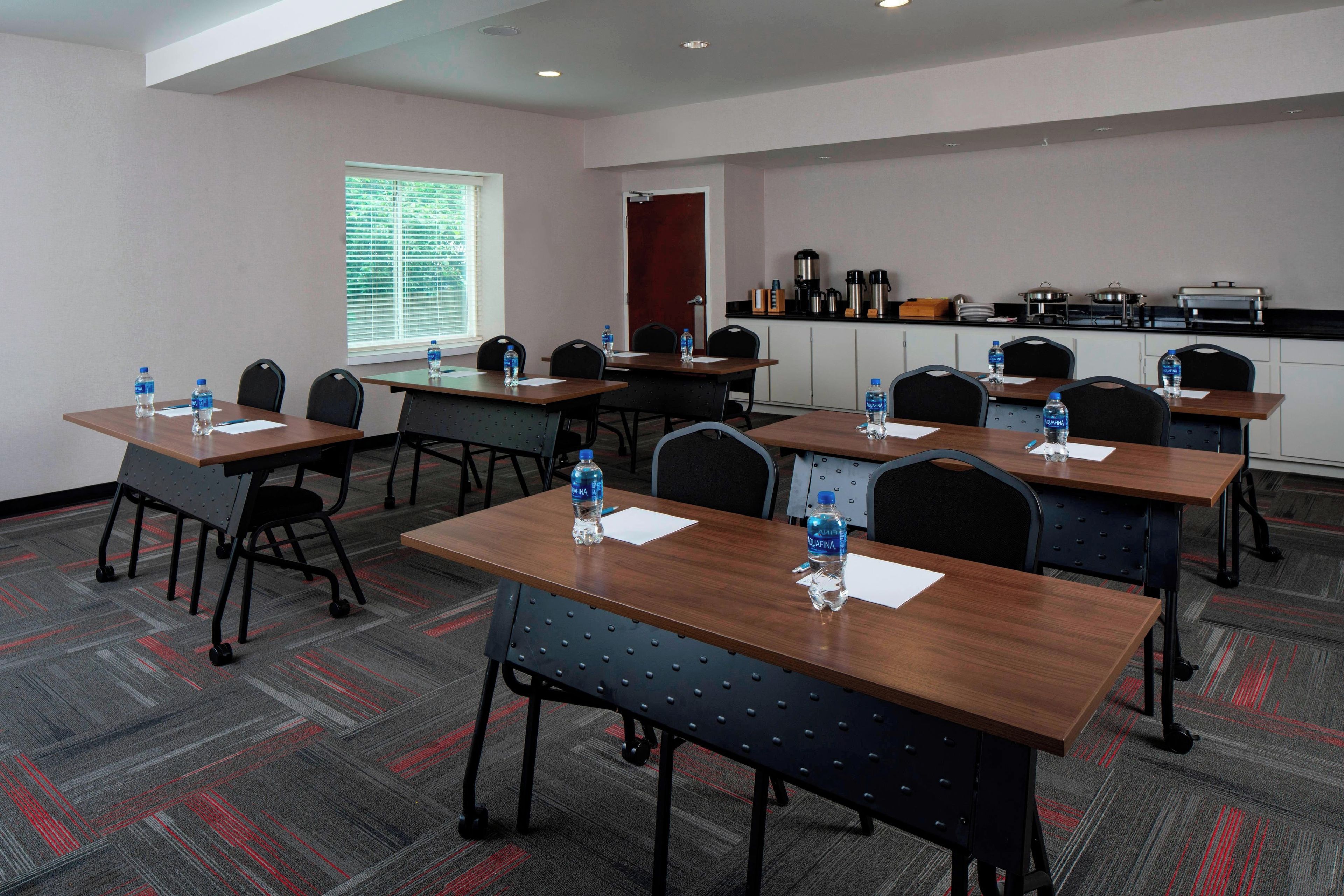A conference room with tables, chairs, water bottles, a window with blinds, a kitchenette, and a door.