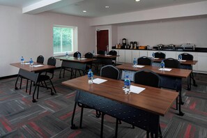 Un salón de conferencias con mesas, sillas, botellas de agua, una ventana con persianas, una kitchenette y una puerta.