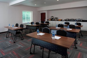Un salón de conferencias con mesas, sillas, botellas de agua, una ventana con persianas, una kitchenette y una puerta.