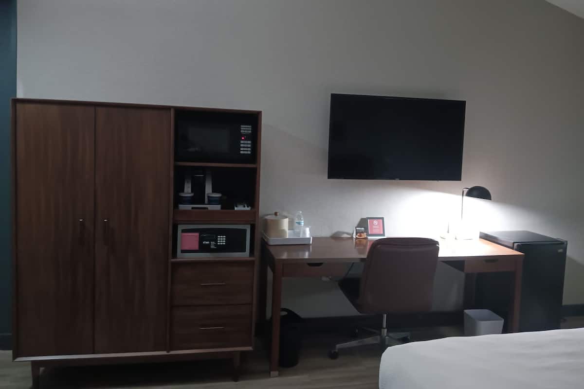 A hotel room with a wooden wardrobe, a desk with a chair, a flat-screen TV, a bedside lamp, and a bed.