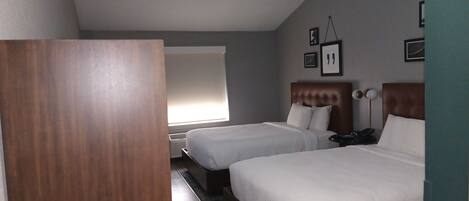 A hotel room with two beds, a window, a bedside table, and a wall-mounted lamp.