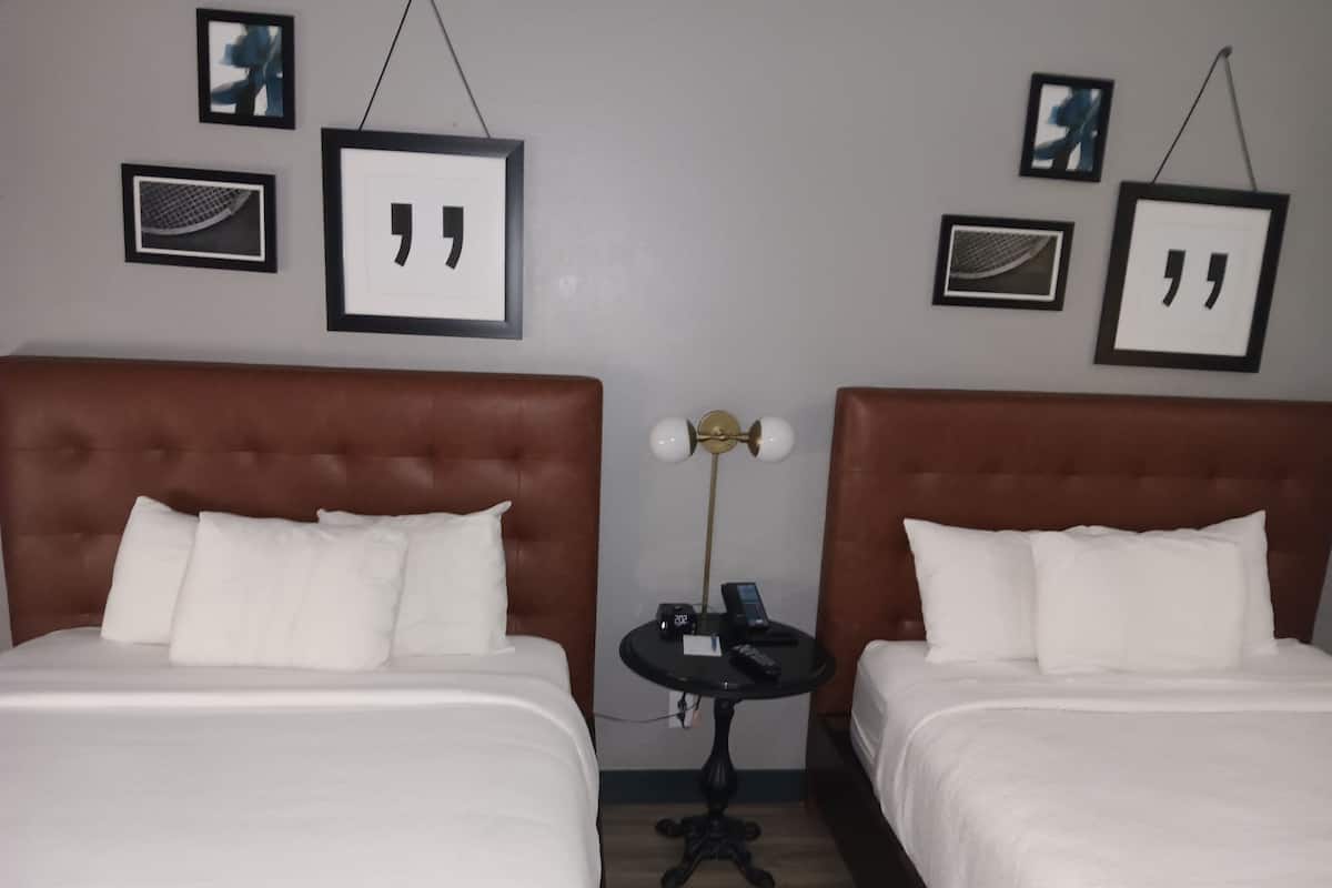 A hotel room with two beds, a small table with a phone and a lamp, and framed quotes on the wall.