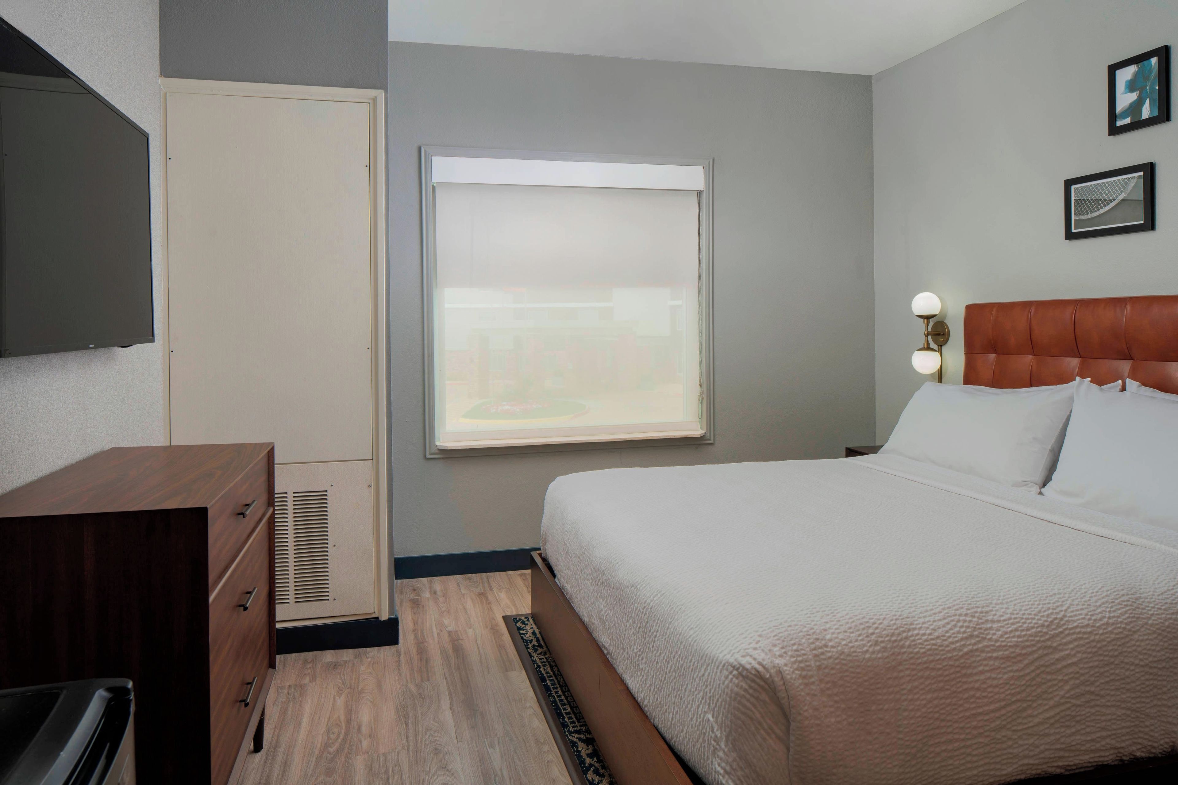 Une chambre d’hôtel comprenant un lit, une commode, une fenêtre avec des stores, un luminaire fixé au mur et un cadre photo.