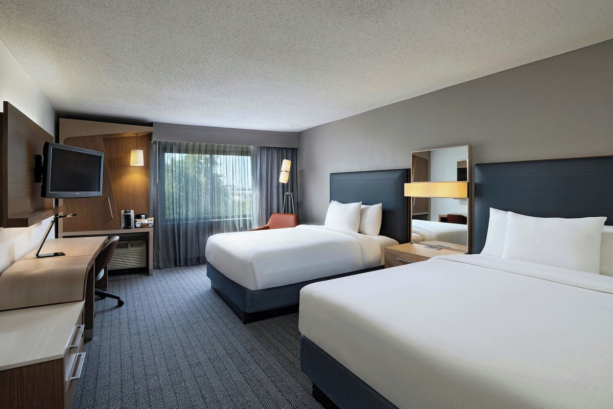 A hotel room with two beds, a desk, a TV, and a window with curtains.