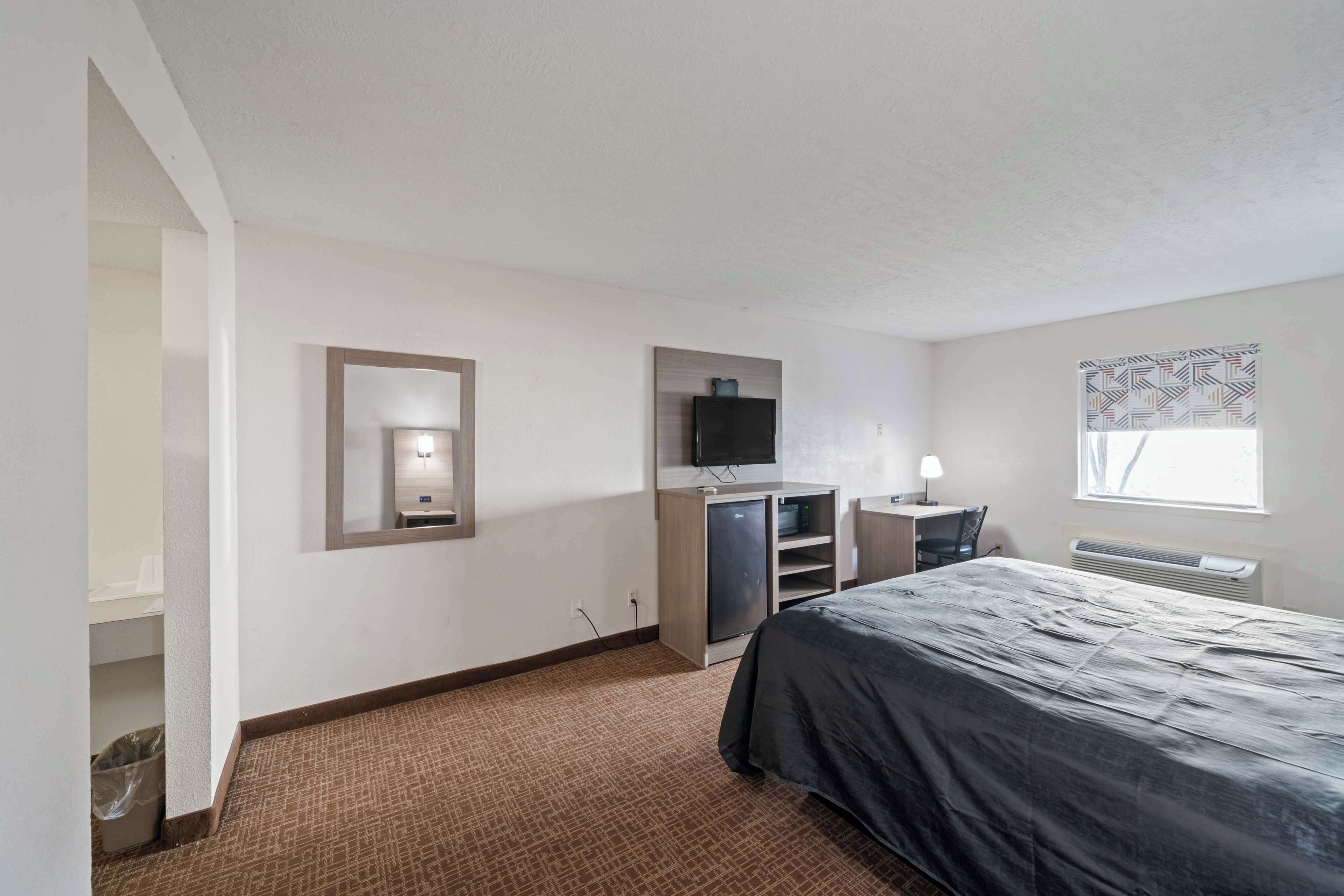 A hotel room with a bed, a desk with a chair, a television, and a window with patterned curtains.