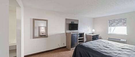 A hotel room with a bed, a desk with a chair, a television, and a window with patterned curtains.