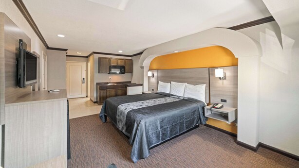 A hotel room with a bed, bedside table, lamp, and a TV mounted on the wall. - Americas Best Value Inn & Suites Brenham (Brenham)