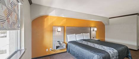Una habitación de hotel con una cama, una mesita de noche con teléfono, una ventana con cortinas estampadas y una lámpara de pared.