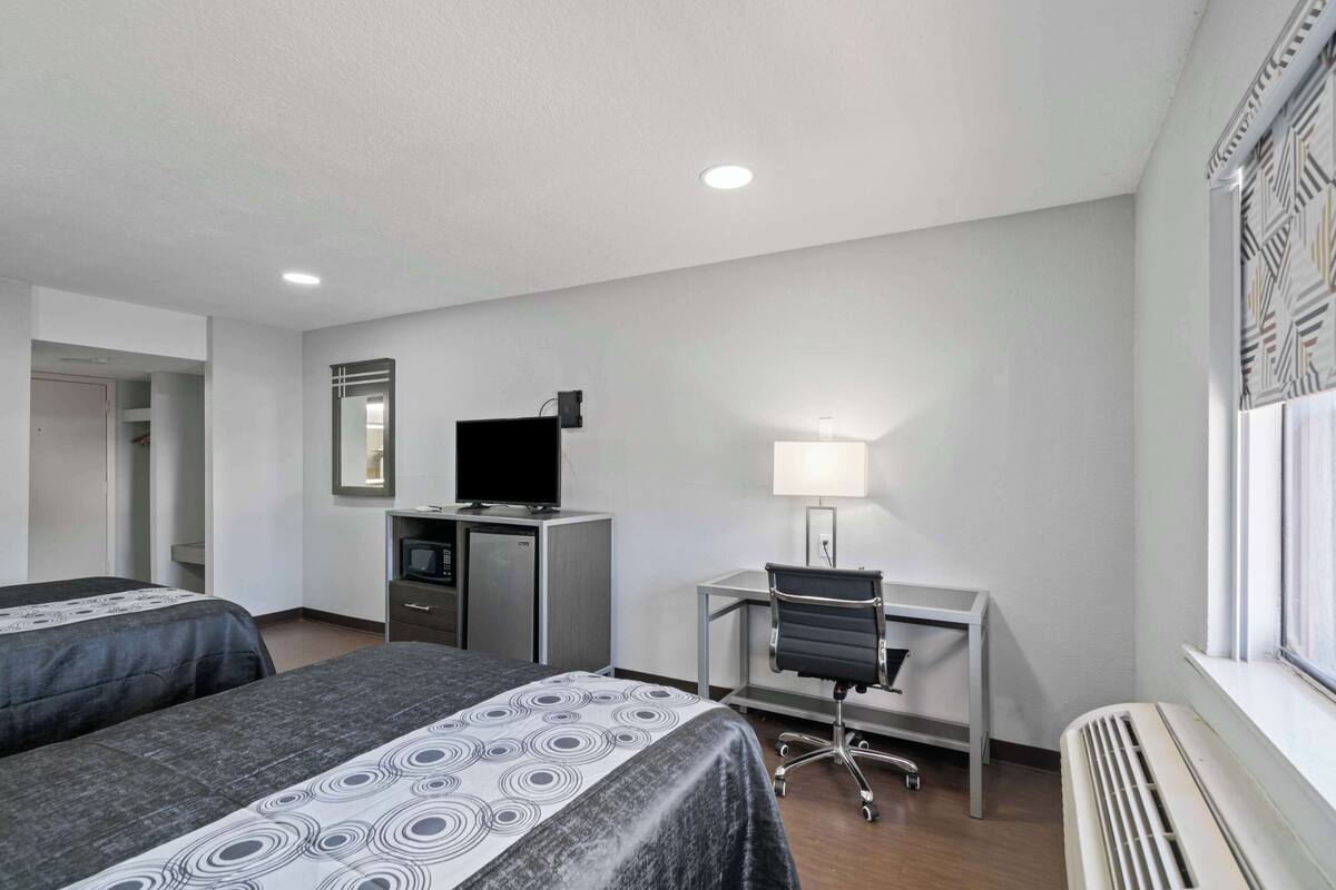 A hotel room with two beds, a desk with a computer and lamp, a TV, and a window with patterned curtains.