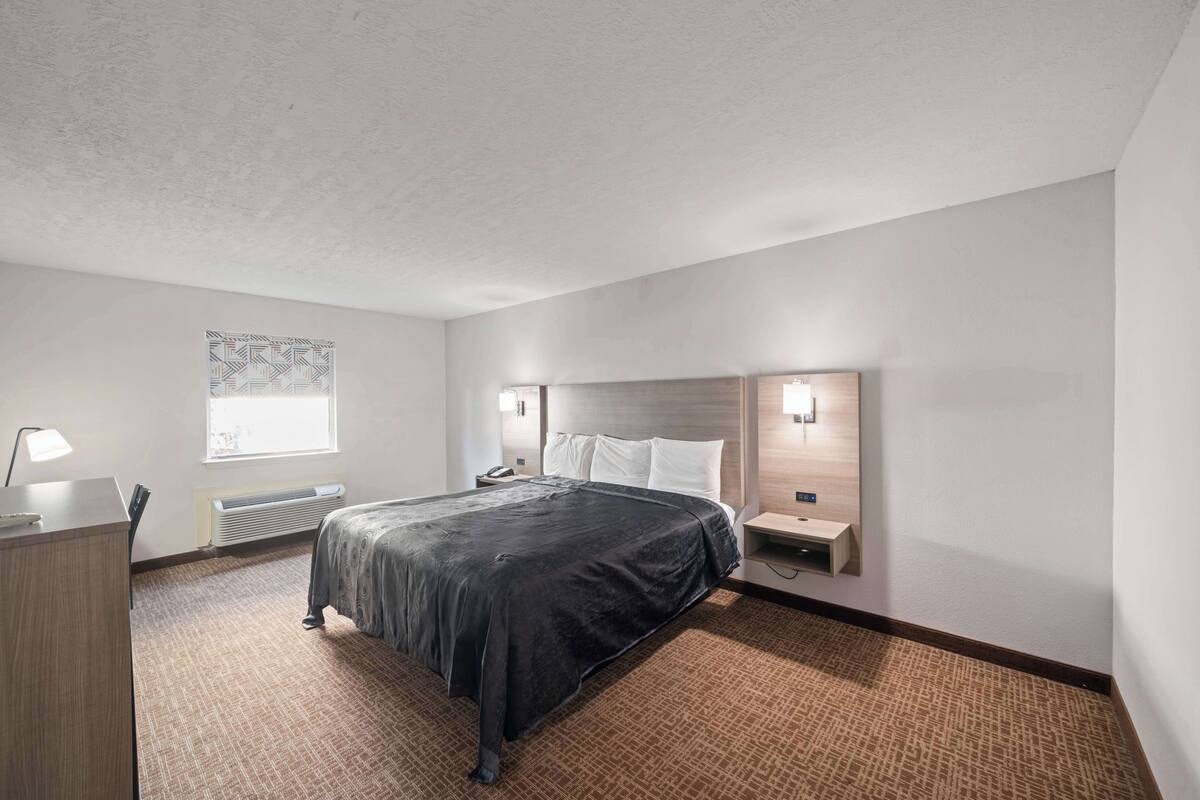 A hotel room with a large bed, a desk with a lamp, a nightstand, a wall-mounted heater, and a window with blinds.