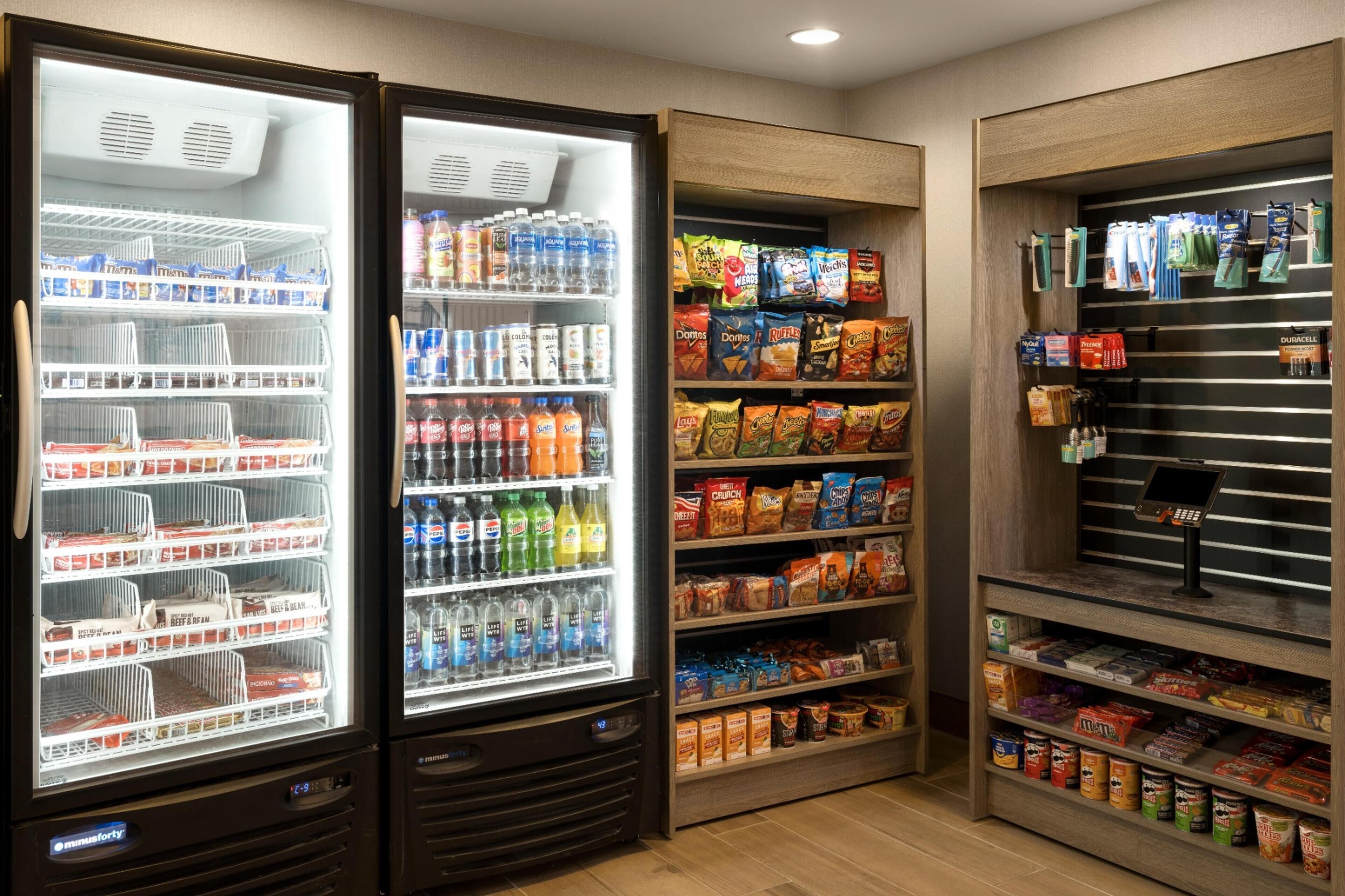 A self-service beverage and snack area with refrigerated drink dispensers, shelves stocked with various beverages, snacks, and packaged goods.