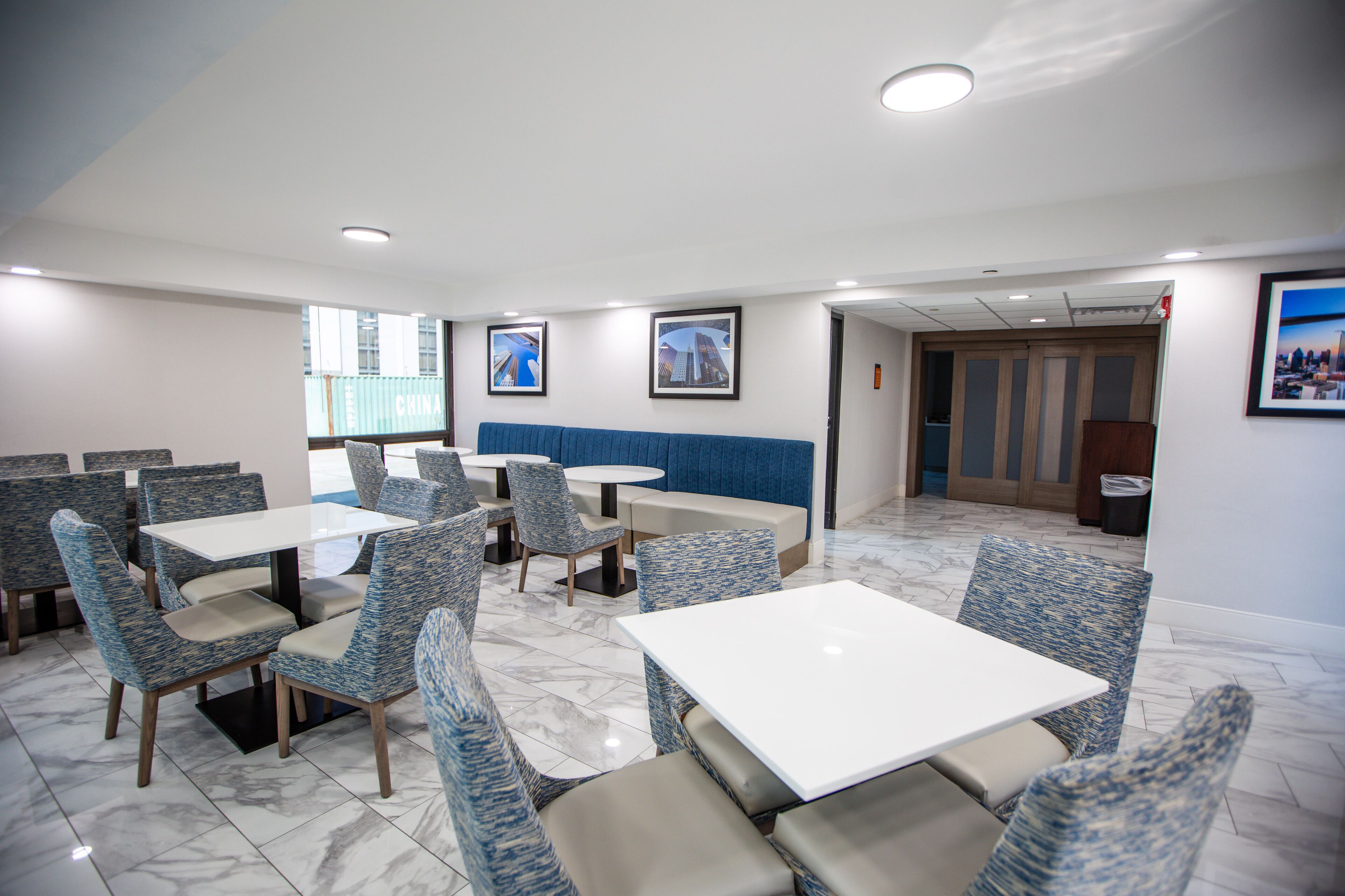 Un espace repas moderne avec des chaises à revêtement bleu et des tables blanches, agrémenté d’un banc rembourré bleu et de tableaux encadrés aux murs.