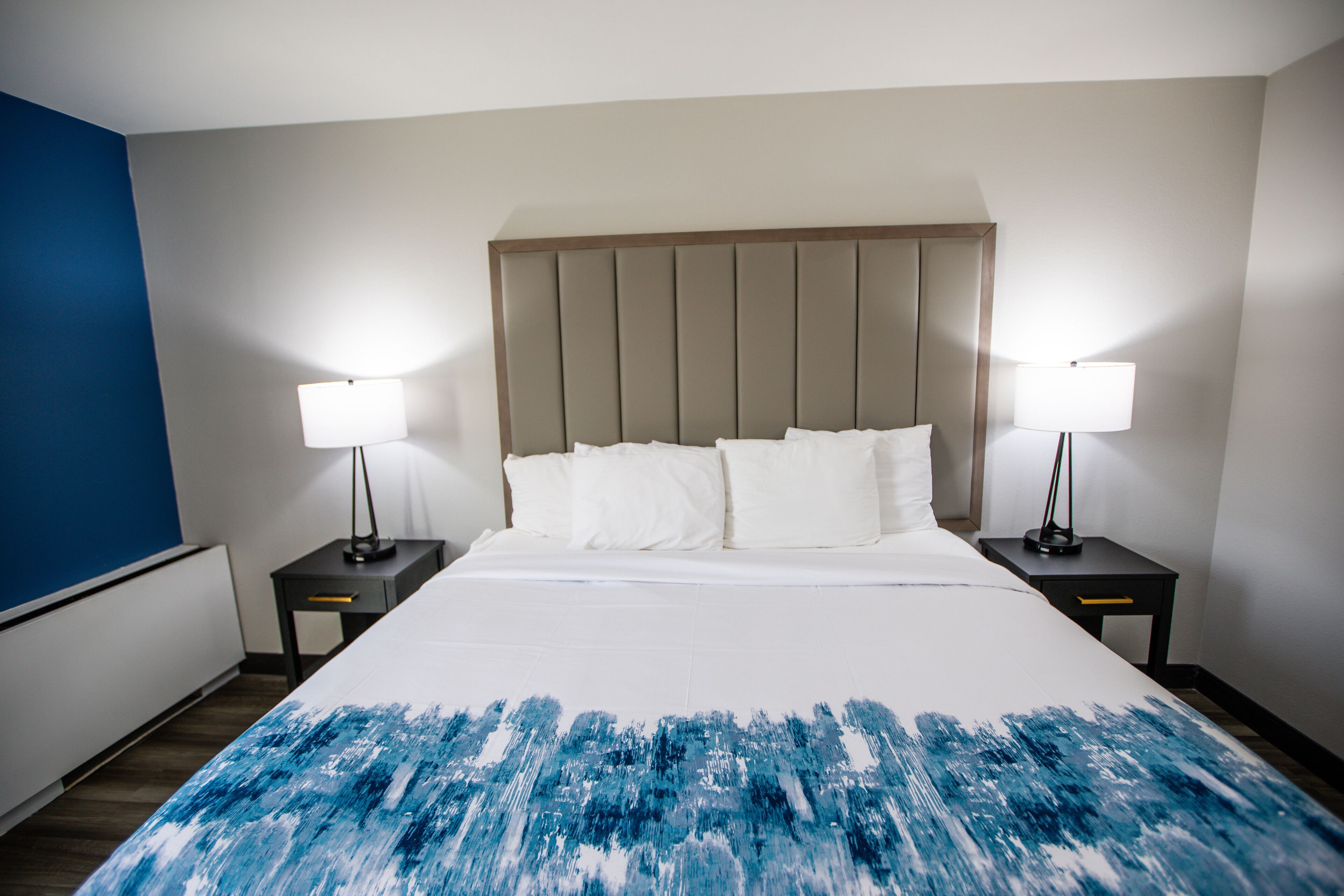 Habitació d'hotel amb un llit gran, dues taules de nit amb làmpades, una paret d'accent blava i un panell de capçal blanc.