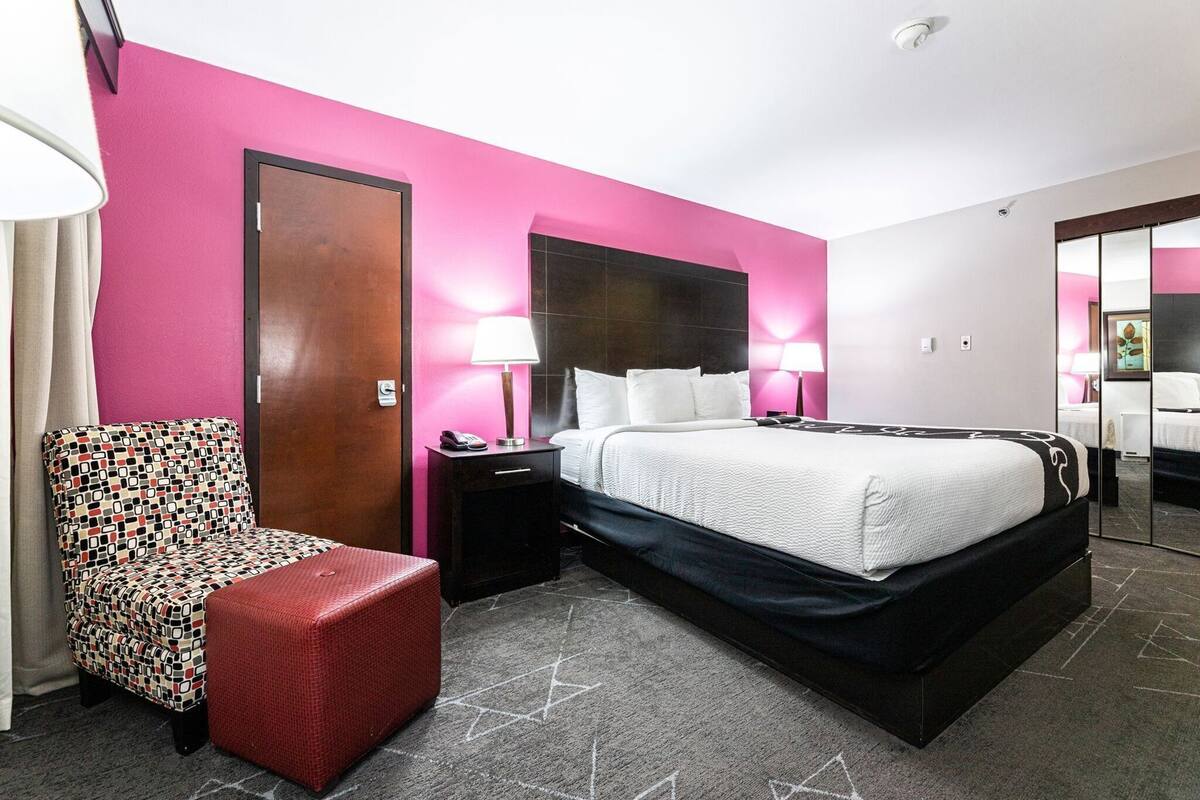 Een hotelkamer met een bed, nachtkastje, stoel en een rode poef.