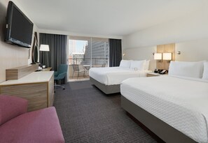 A hotel room with two beds, a desk, a TV, a chair, and a balcony with a city view.