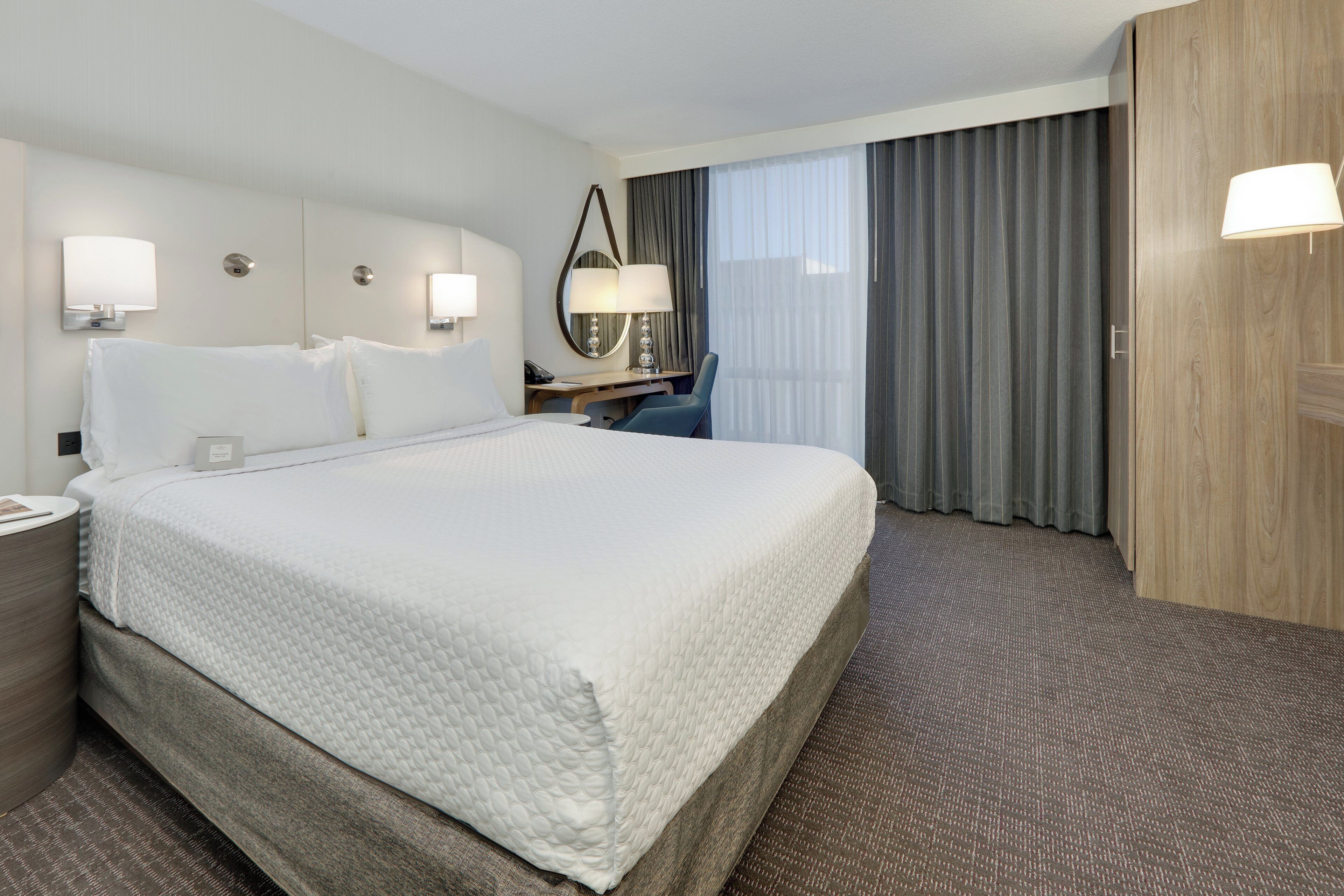 Une chambre d’hôtel avec un grand lit, un bureau, une chaise, un miroir et une fenêtre avec des rideaux.