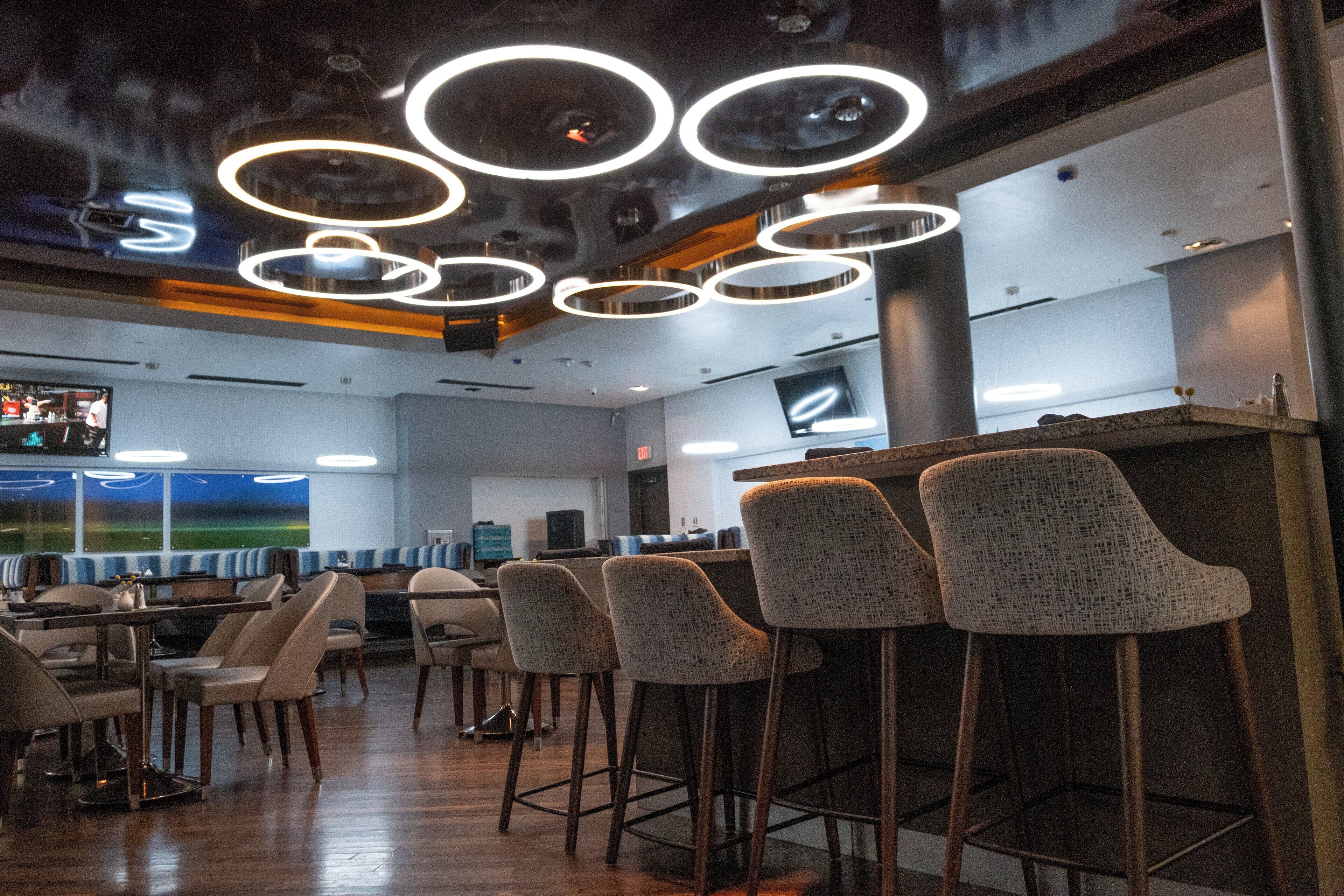 A modern bar area with multiple seating options, including high stools and bar chairs, and a television screen mounted on the wall.