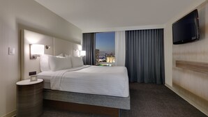 A hotel room with a bed, a bedside table, a TV mounted on the wall, and a view of the cityscape at night. - Crowne Plaza Dallas Downtown by IHG (Dallas)