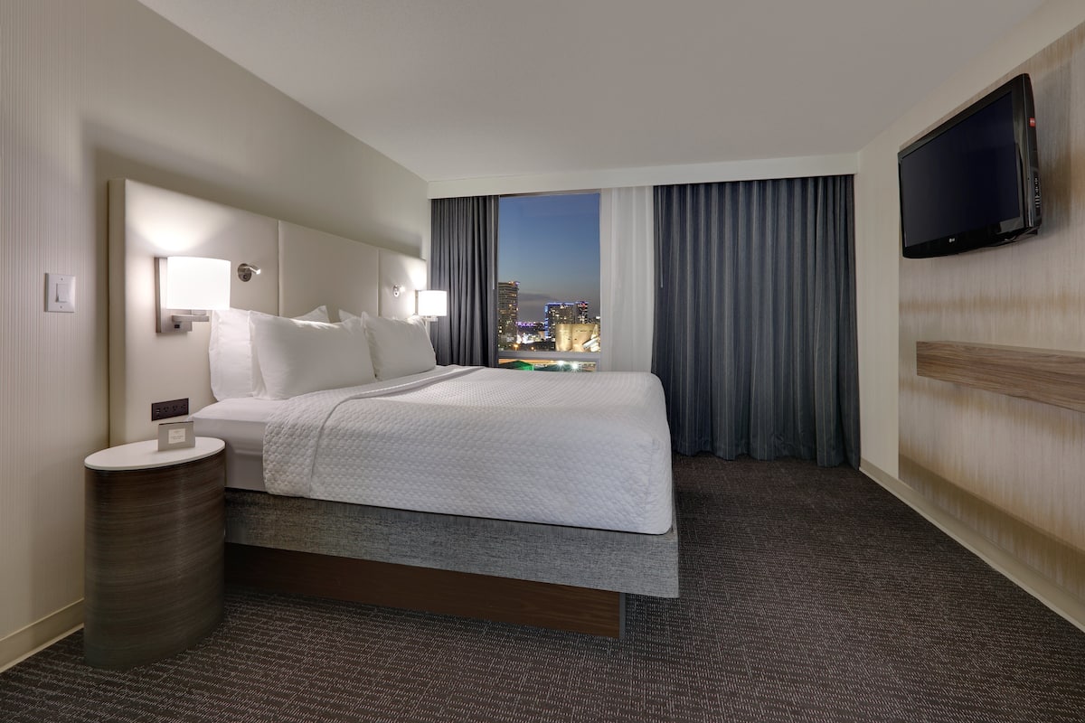 A hotel room with a bed, a bedside table, a TV mounted on the wall, and a view of the cityscape at night.