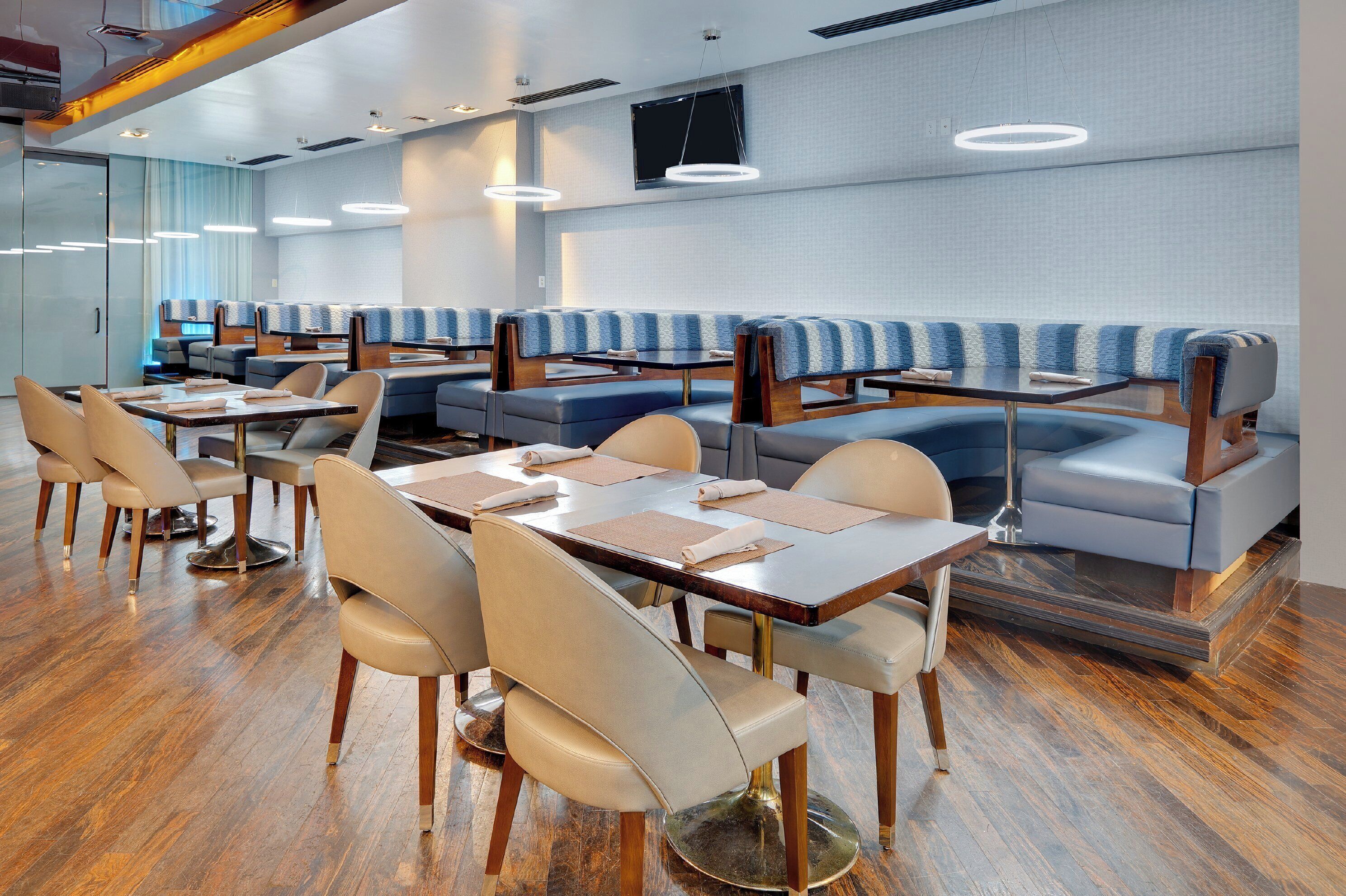 A modern restaurant with booths and tables, wooden flooring, and a large flat-screen TV on the wall.