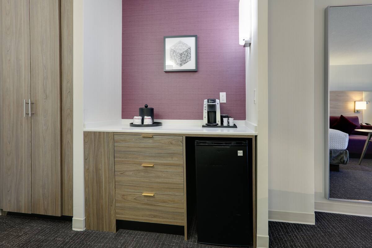A compact hotel room with a wooden wardrobe, a small desk with a coffee machine, and a compact refrigerator.