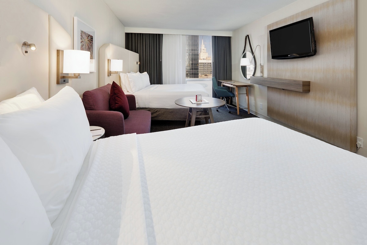 A hotel room with a large bed, a desk with a chair, a small table, and a TV mounted on the wall.