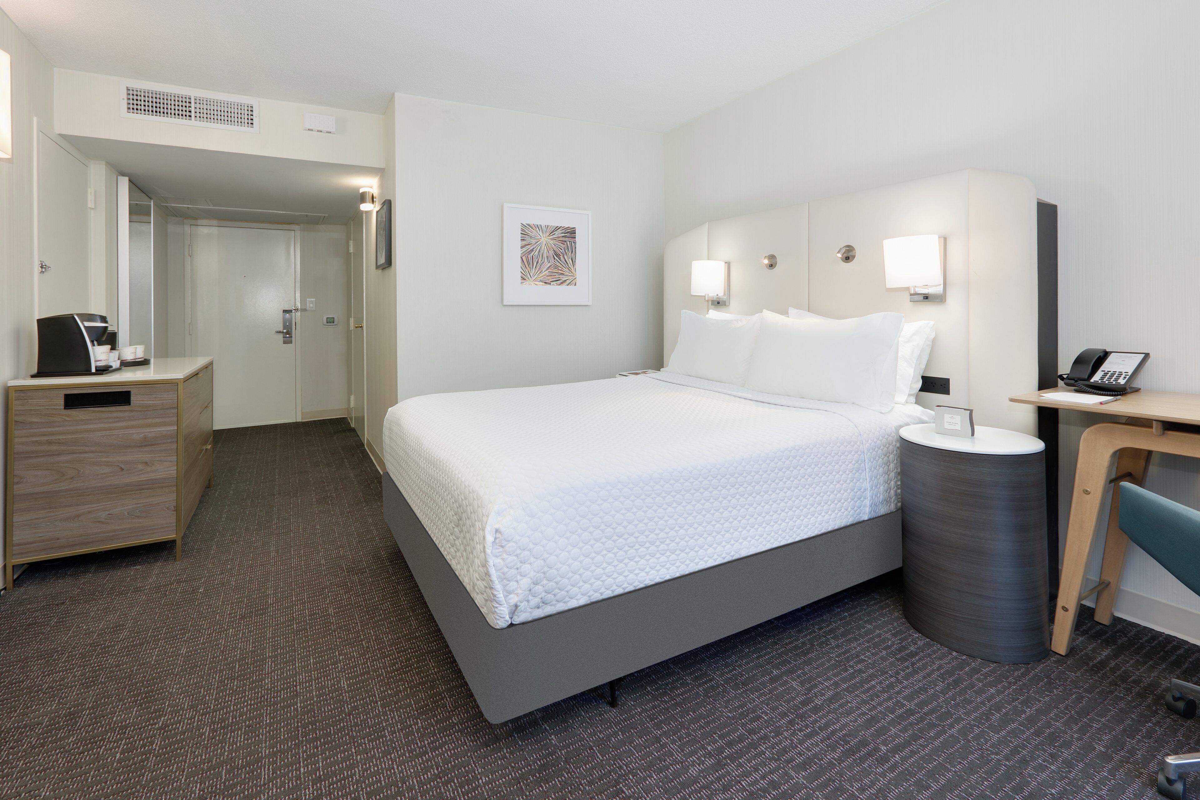 A hotel room with a bed, a desk, a chair, a telephone, and a waste bin.
