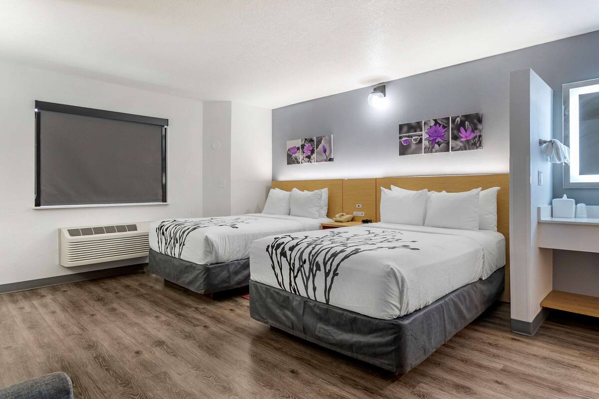A hotel room with two beds, a large window, a wall-mounted air conditioner, and a bathroom with a mirror and towel rack.