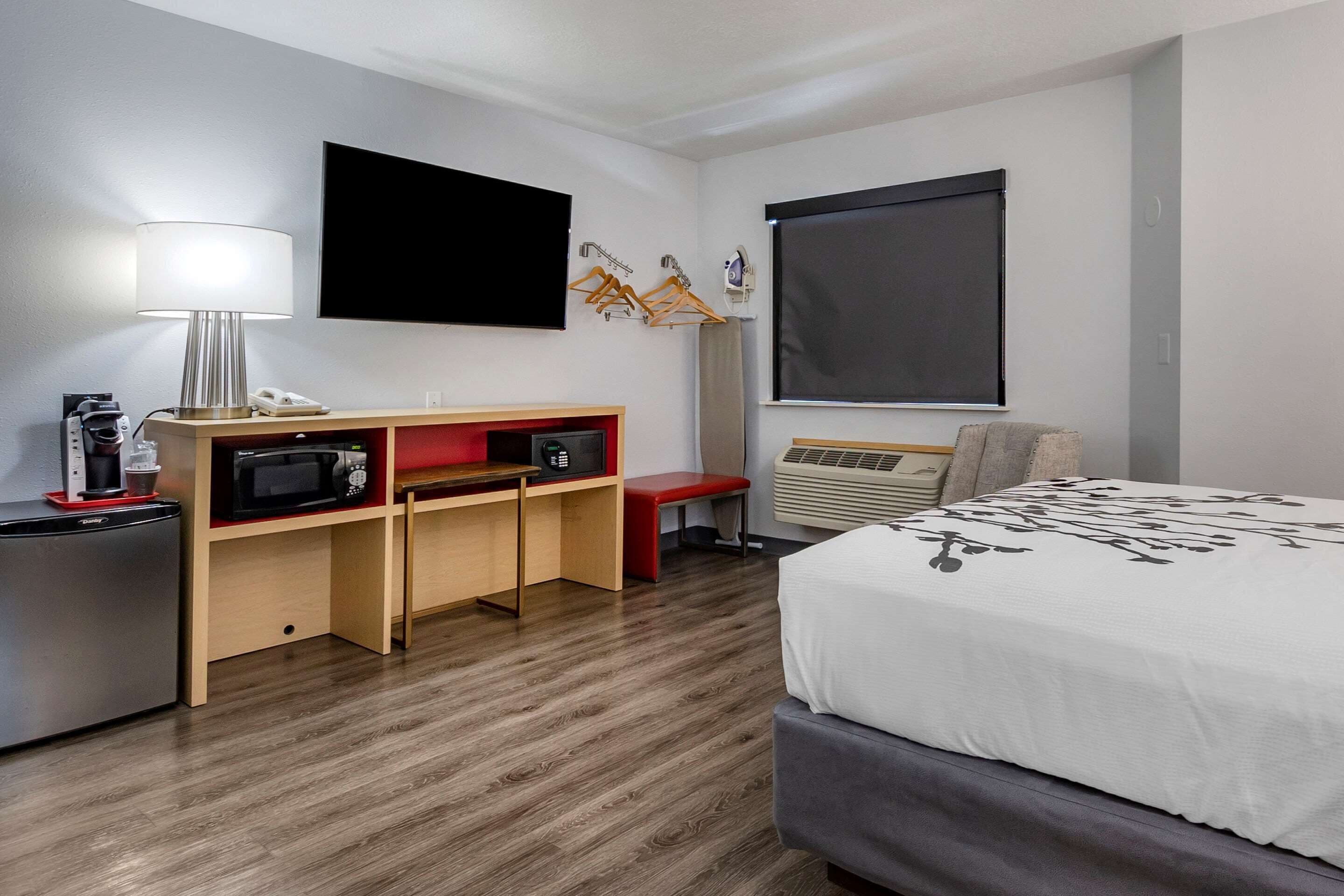Habitación de hotel con cama, escritorio con lámpara, microondas, televisor y ventana con persiana.