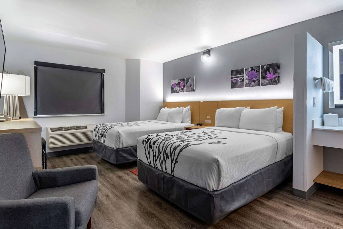 A hotel room with two beds, a desk, a chair, a flat-screen TV, and artwork on the wall.