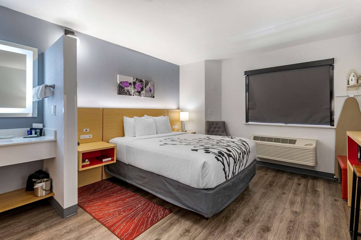A hotel room with a bed, bedside table, nightstand, wall-mounted heater, and a window with a blind.