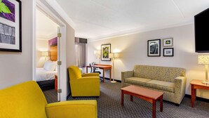 A hotel room with a bed, a yellow armchair, a patterned sofa, a small table, and a TV.