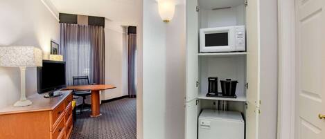 A hotel room with a wooden desk, a flat-screen TV, a lamp, a microwave, a mini-fridge, and a cabinet with kitchen items.