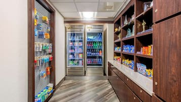Un corridoio stretto con due distributori automatici di bevande refrigerate, scaffali pieni di snack e bevande, e un armadietto in legno con diversi oggetti.